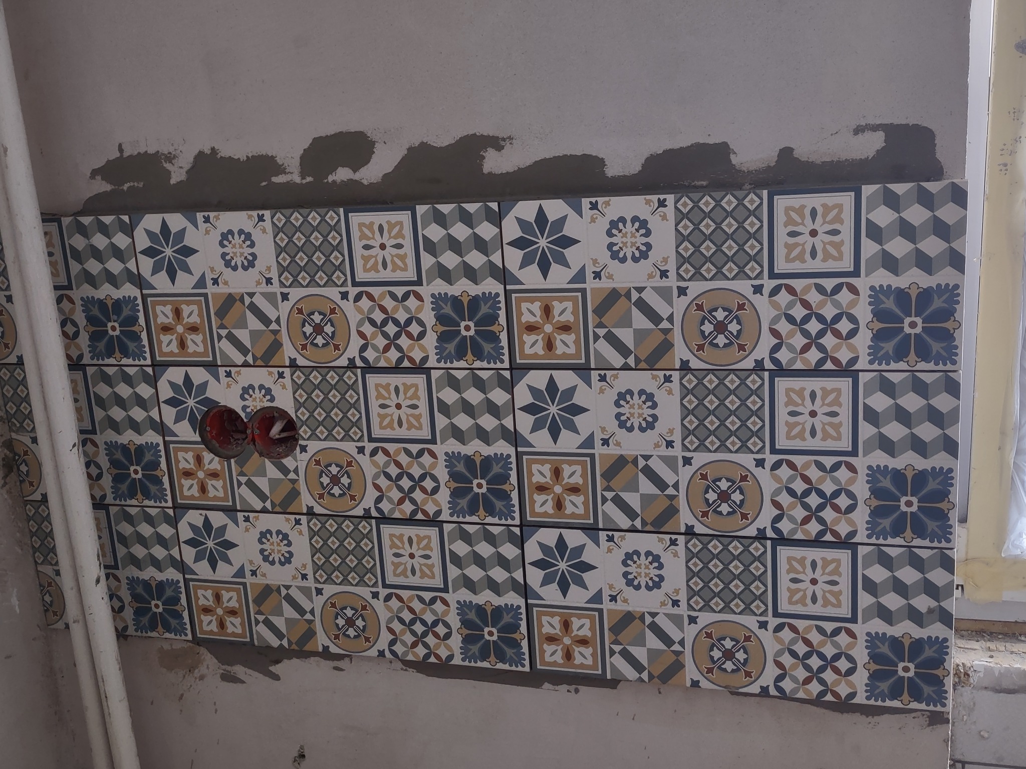 Fragment ściany z ułożonymi, dekoracyjnymi płytkami ceramicznymi w stylu patchwork, widoczne puszki elektryczne bez osprzętu i ślady zaprawy na ścianie.
