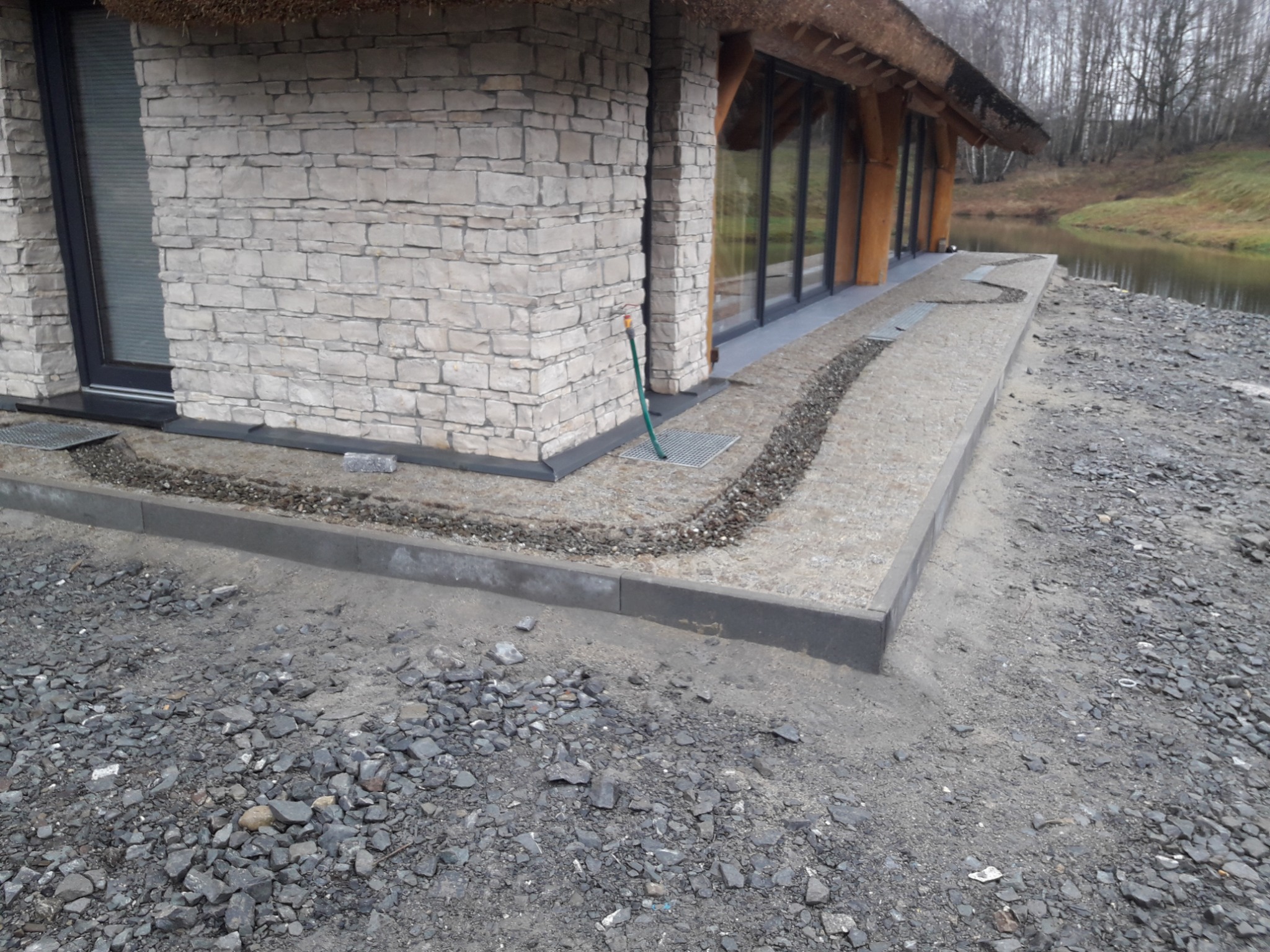 Wykończona elewacja budynku z kamienia z widocznym fragmentem dachu krytego strzechą, obok ułożona kostka brukowa i ozdobny pas z drobnych kamieni, w tle zbiornik wodny i drzewa.