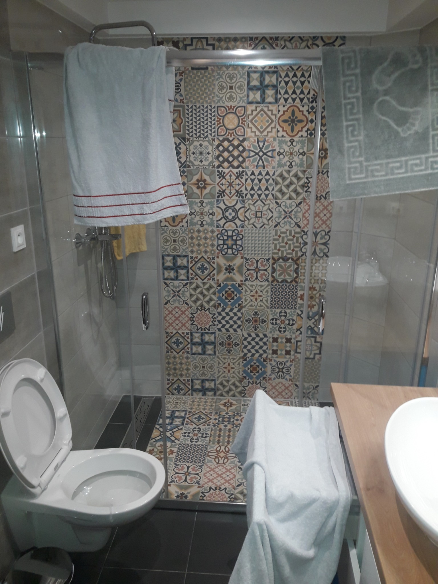 Ukończona łazienka z kabiną prysznicową wyłożoną mozaiką w stylu patchwork, białą umywalką nablatową i toaletą z otwartą klapą.