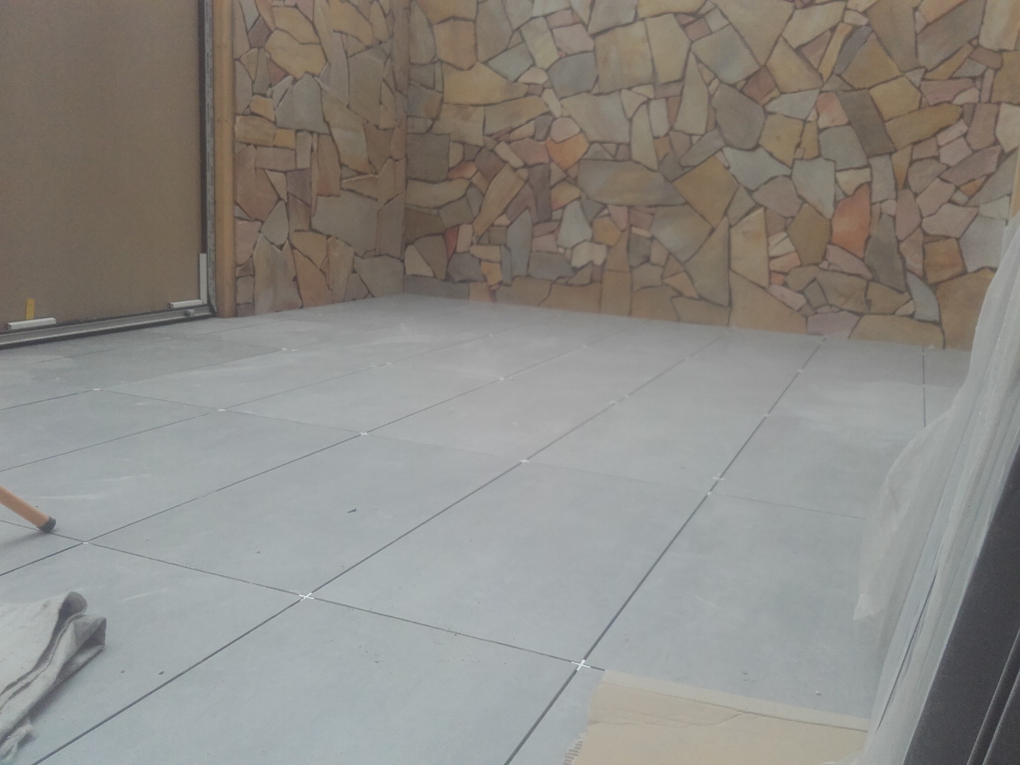 Ułożona szara podłoga z płytek ceramicznych z krzyżykami dystansowymi, ściana wyłożona kamieniem naturalnym o nieregularnych kształtach i ciepłych barwach, widoczny fragment poziomicy i materiału...