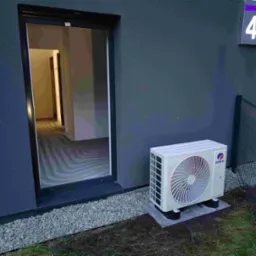 Zewnętrzna jednostka klimatyzacji GREE zamontowana na betonowych podstawach przy szarej ścianie budynku z numerem 43, widok zza ogrodzenia.