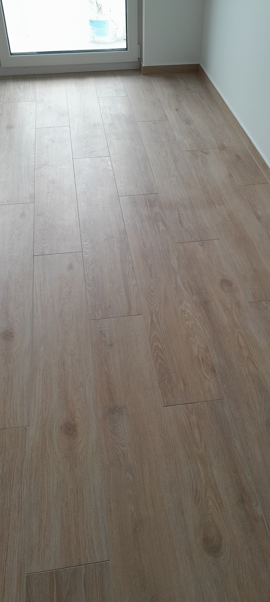 Ułożona podłoga z paneli imitujących jasne drewno, widok z góry, przy ścianie biała listwa przypodłogowa, w tle okno.