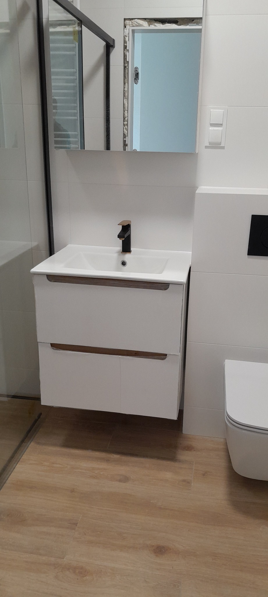 Biała szafka pod umywalkę z czarną baterią, wisząca toaleta i kabina prysznicowa w wyremontowanej łazience z panelami podłogowymi.