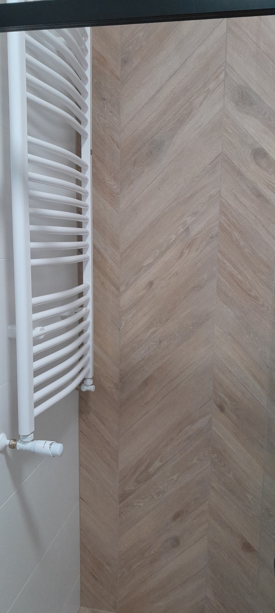 Biały, drabinkowy grzejnik łazienkowy na tle ściany wyłożonej jasnymi, drewnopodobnymi płytkami ułożonymi w jodełkę.