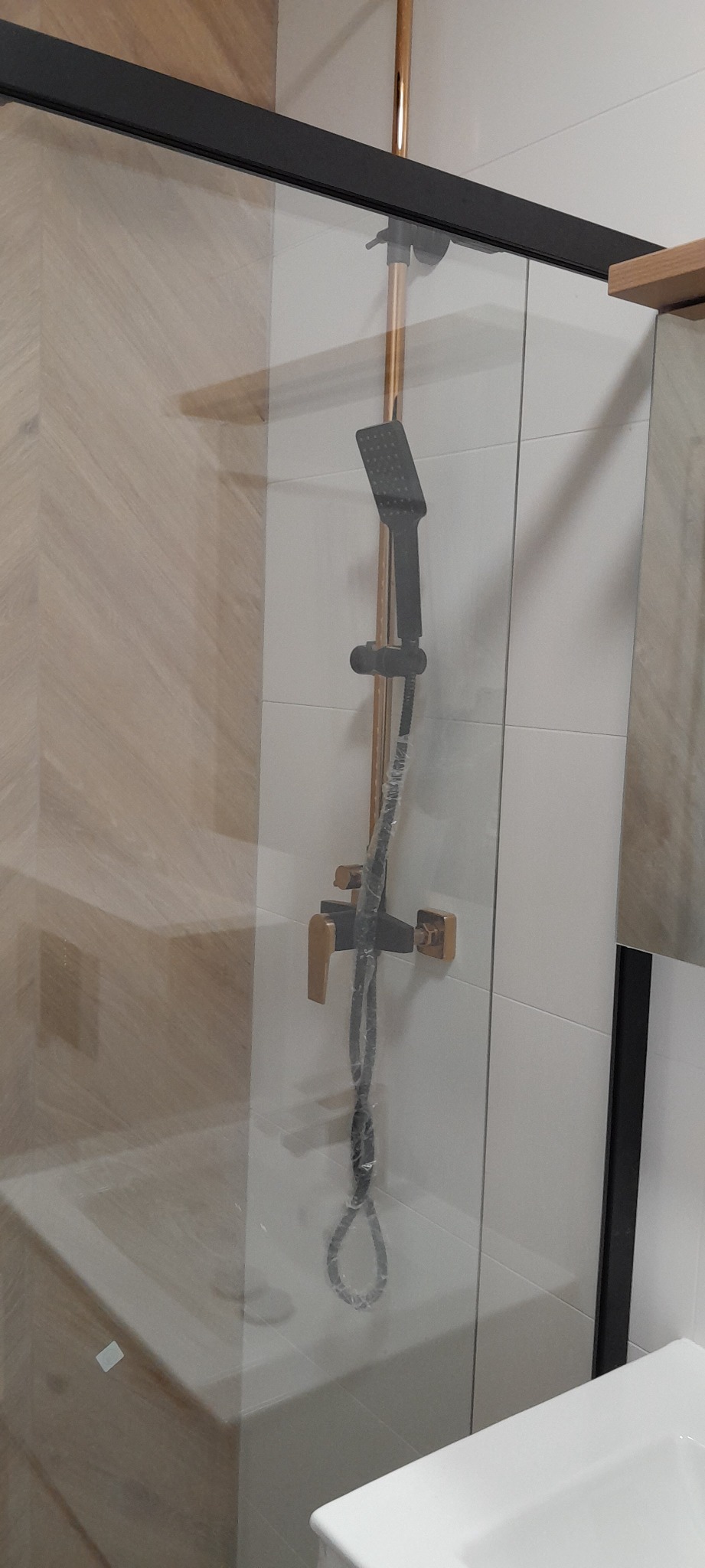Nowoczesna kabina prysznicowa z czarną ramą i złotą armaturą, jeszcze w folii ochronnej, na tle białych i drewnopodobnych płytek.