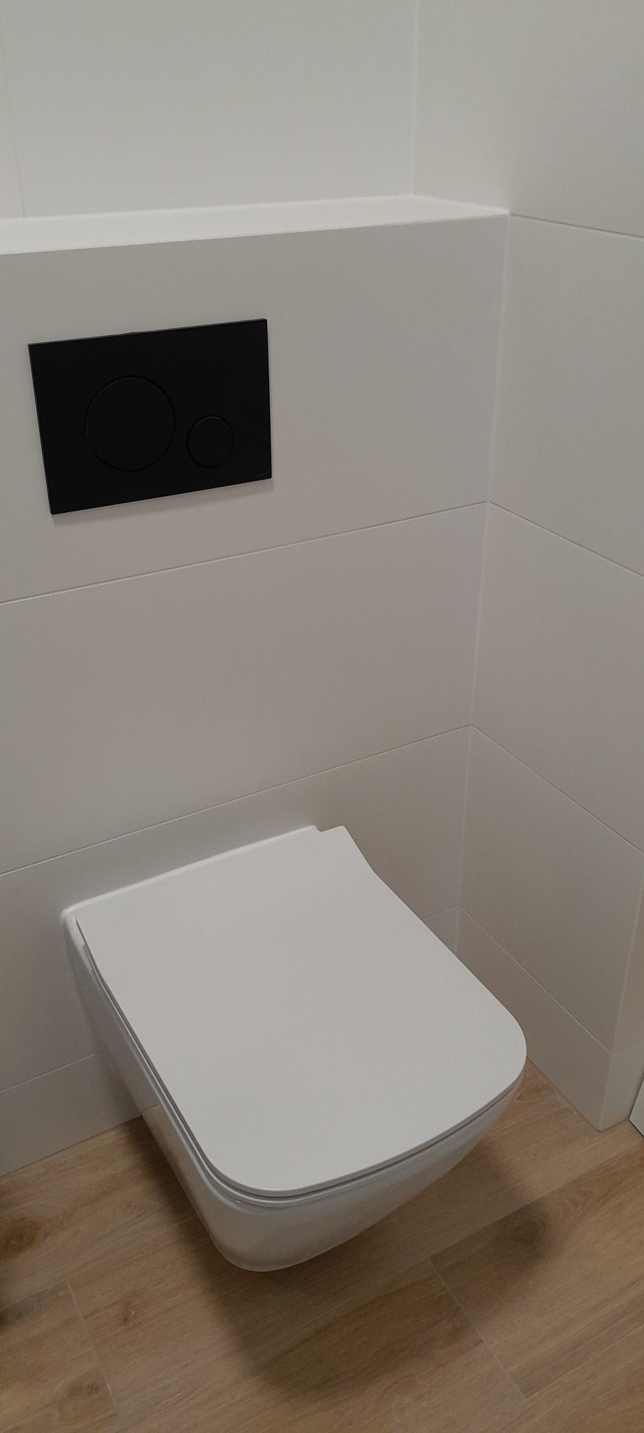Nowoczesna, podwieszana toaleta z czarnym panelem spłukującym na białych, poziomych płytkach i drewnopodobną podłogą.