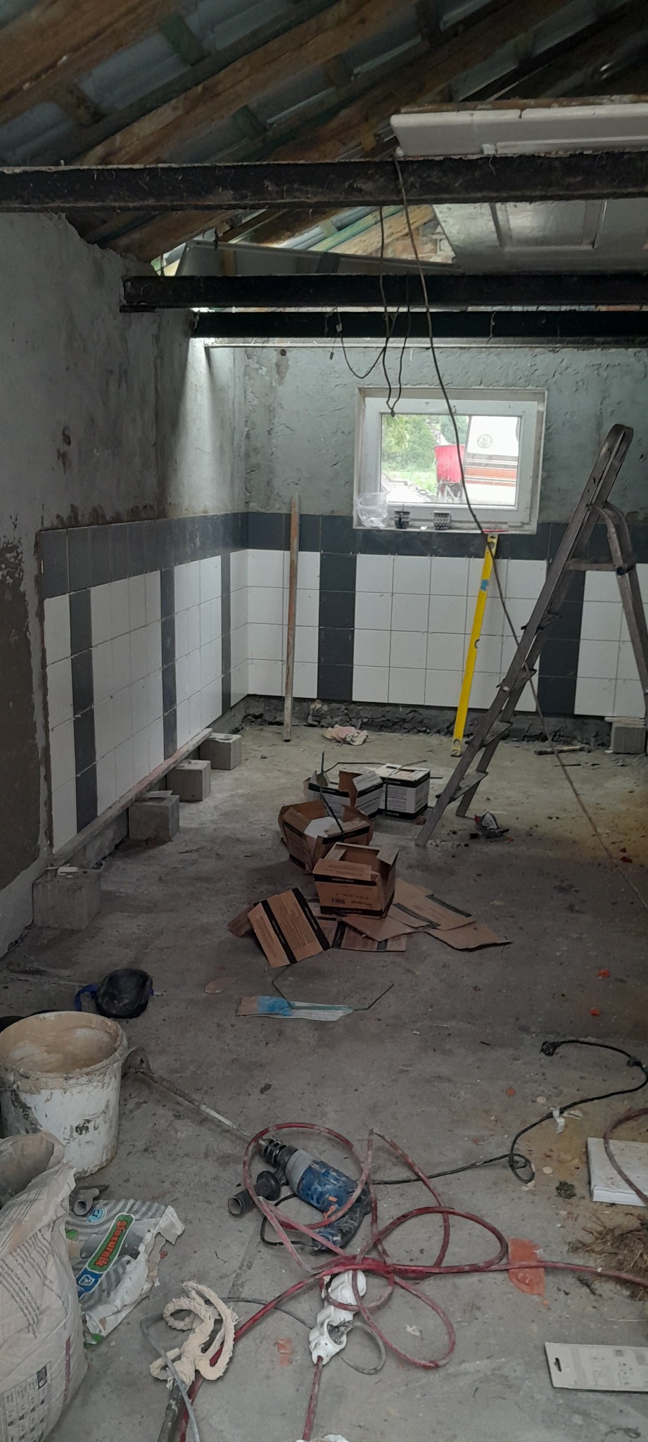 Wnętrze w trakcie remontu, częściowo wyłożone płytkami w biało-szare pasy, widoczne narzędzia glazurnicze, poziomica oparta o ścianę, surowy beton na podłodze i odsłonięta konstrukcja dachu.