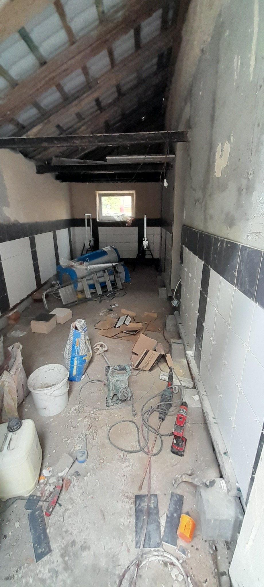 Wnętrze w trakcie układania płytek, widoczne narzędzia, mieszadło, poziomica, wiadro oraz worki z zaprawą, ściany częściowo wyłożone białymi i czarnymi płytkami.