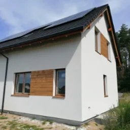 Nowoczesny dom jednorodzinny z białą elewacją i drewnianymi akcentami wokół okien, panele solarne na dachu, widok z zewnątrz w słoneczny dzień.