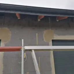 Fragment ściany budynku z szarym styropianem, częściowo pokrytym warstwą zaprawy, widoczne rusztowanie i okna z roletami zewnętrznymi.