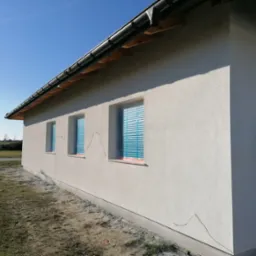 Świeżo otynkowana elewacja domu jednorodzinnego z zabezpieczonymi oknami folią, widoczne ślady po przewodach elektrycznych wystających ze ściany.