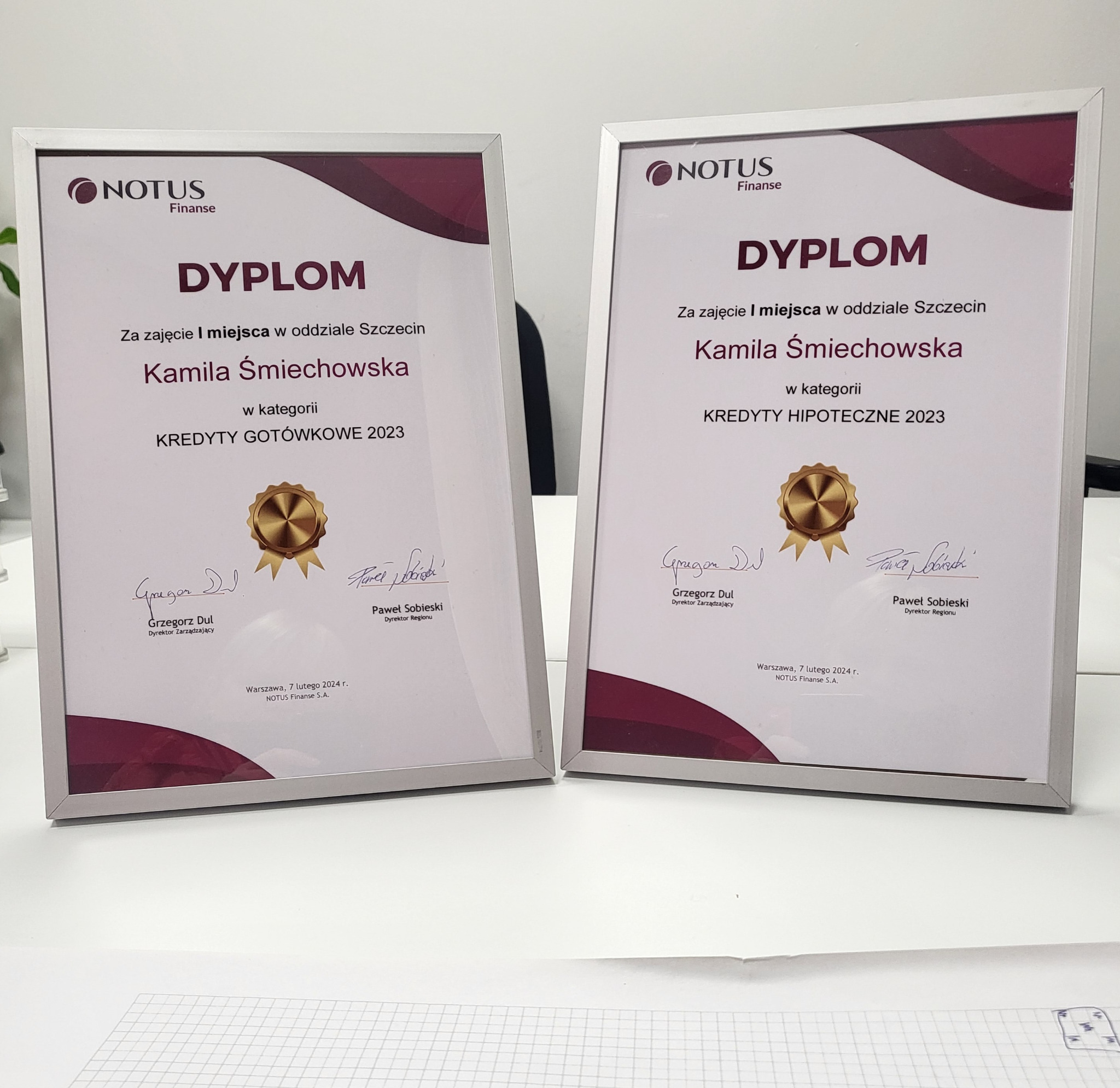 Dwa dyplomy w srebrnych ramkach na białym biurku, jeden za kredyty gotówkowe, drugi za kredyty hipoteczne, oba dla Kamili Śmiechowskiej z firmy Notus Finanse, za pierwsze miejsce w oddziale Szczecin.