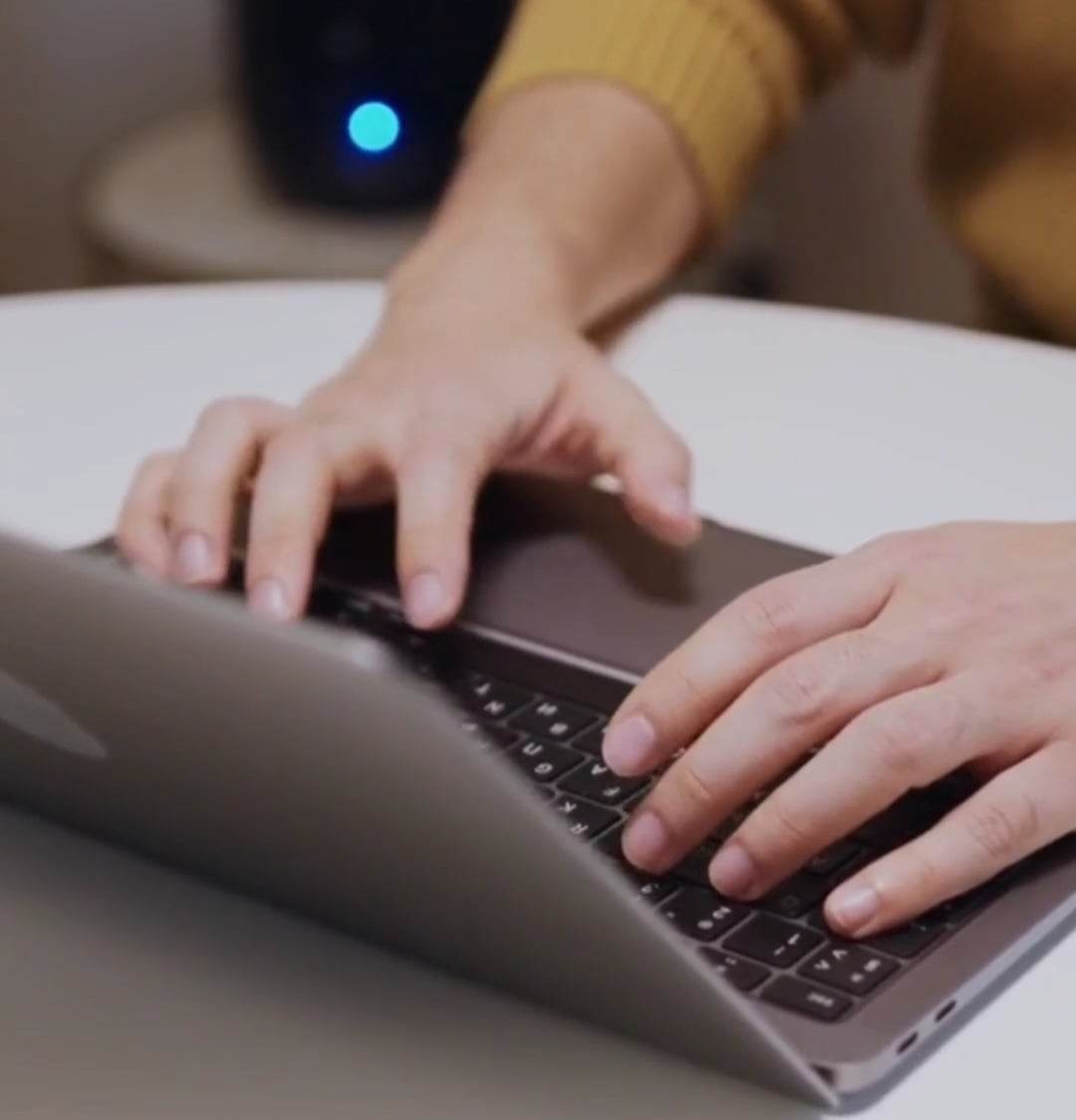 Dłonie mężczyzny piszącego na laptopie z podświetlaną klawiaturą; w tle widoczny router z niebieską diodą.