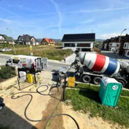 Betoniarka z czerwoną, białą i niebieską spiralą na tle budynków mieszkalnych i błękitnego nieba z chmurami, obok zaparkowany bus z reklamą firmy budowlanej i toaleta przenośna.