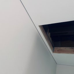 ECOBUD - Biały sufit z otworem włazowym na strych, widoczne drewniane elementy konstrukcyjne i ocieplenie. Minimalistyczne wnętrze, kątowe ujęcie.