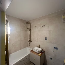 ECOBUD - Wykończona łazienka z beżowymi płytkami, białą wanną z dekoracyjnym wzorem i umywalką na szafce. Widoczne elementy instalacji elektrycznej i hydraulicznej.