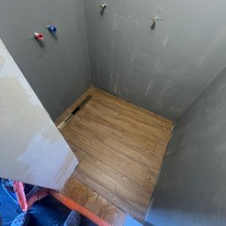 ECOBUD - Szare ściany łazienki z instalacją wodną, drewnopodobne płytki podłogowe z odpływem liniowym. Widoczne narzędzia i fragment osoby w obuwiu roboczym.