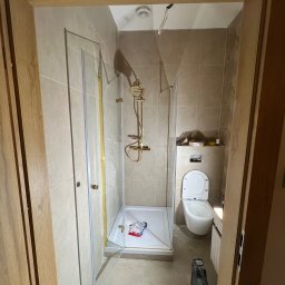 ECOBUD - Wykończona łazienka z kabiną prysznicową ze złotymi elementami, toaletą i beżowymi płytkami. Widok zza drewnianej futryny drzwi.