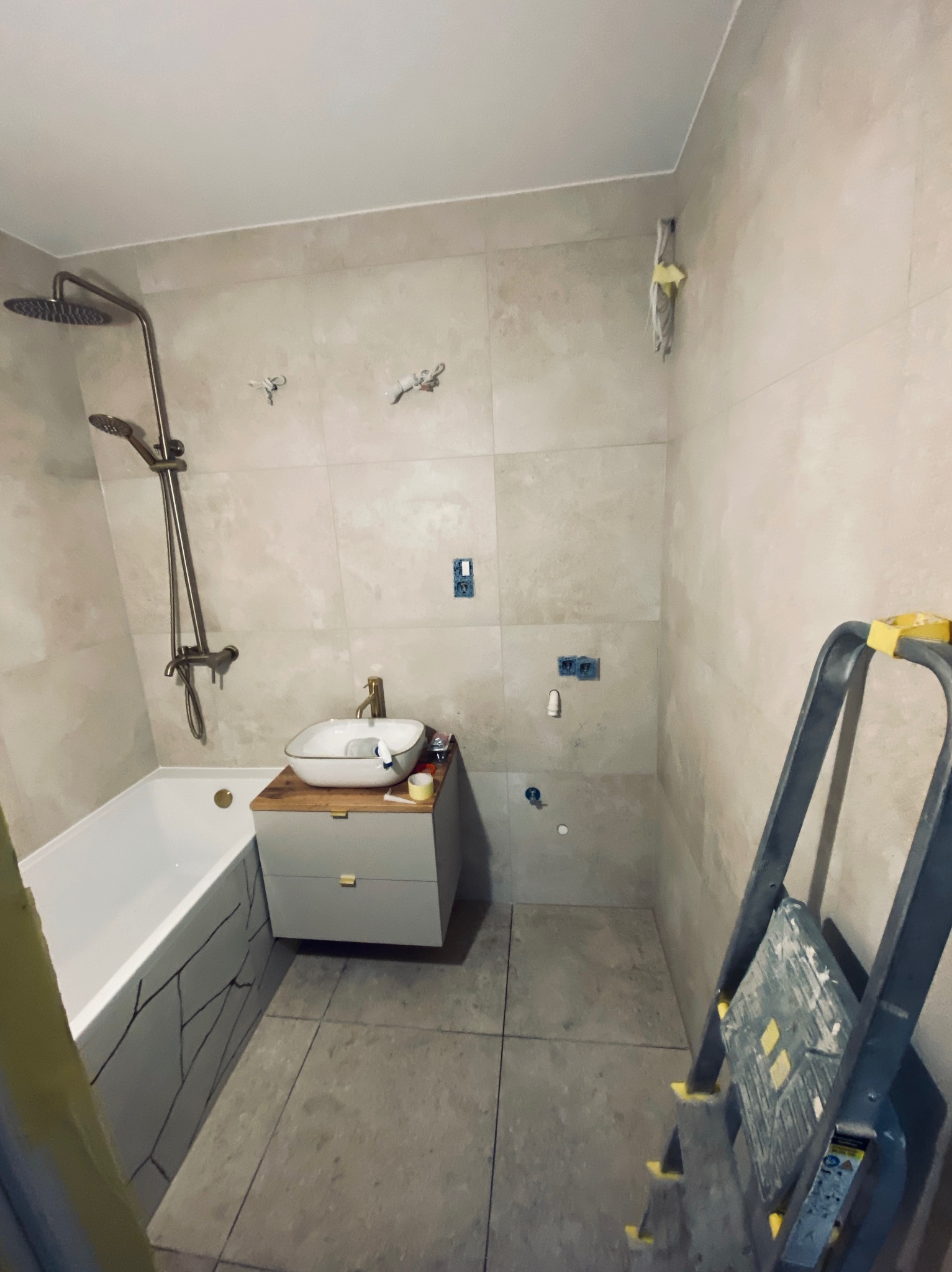 Wykończona łazienka z wanną, umywalką na szafce z drewnianym blatem, baterią w kolorze złota i drabina malarska, płytki na ścianach i podłodze.