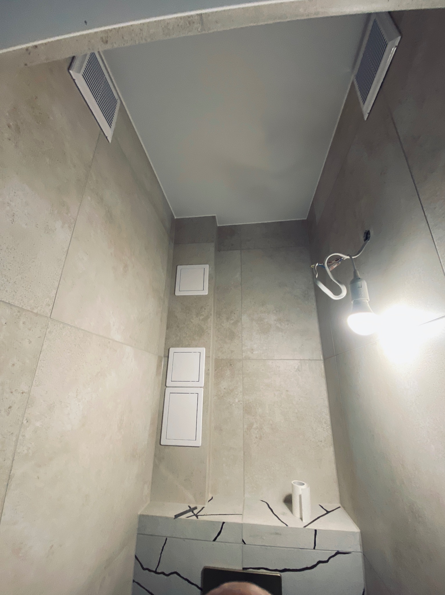 Wykończona łazienka z beżowymi płytkami, białym sufitem podwieszanym i kratkami wentylacyjnymi. Widoczne białe drzwiczki rewizyjne i prowizoryczne oświetlenie.