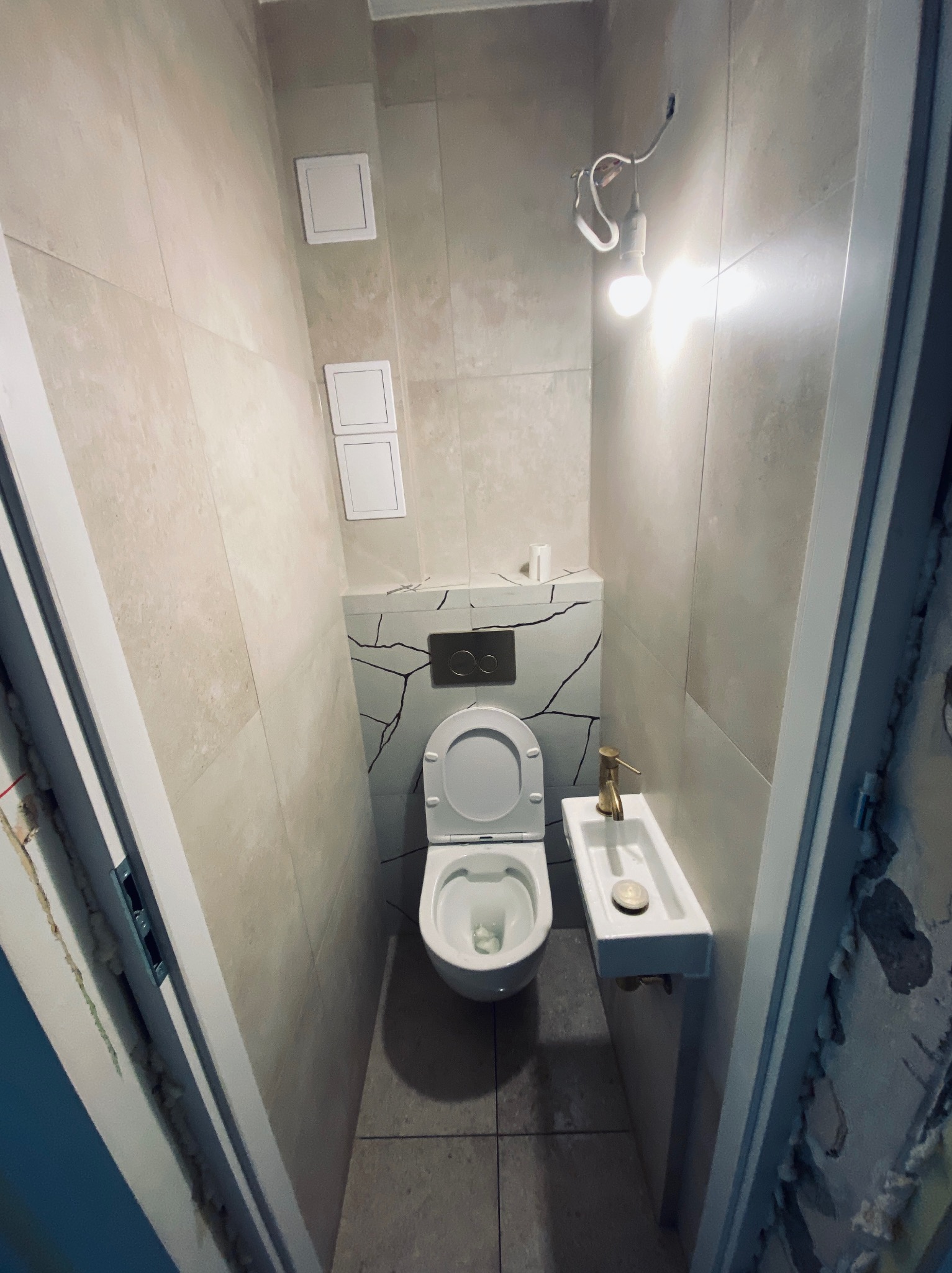 Wąska toaleta z beżowymi płytkami, umywalką z złotą armaturą i motywem pęknięć na ścianie za toaletą. Widoczna żarówka na kablu i niedokończone wykończenie przy futrynie.