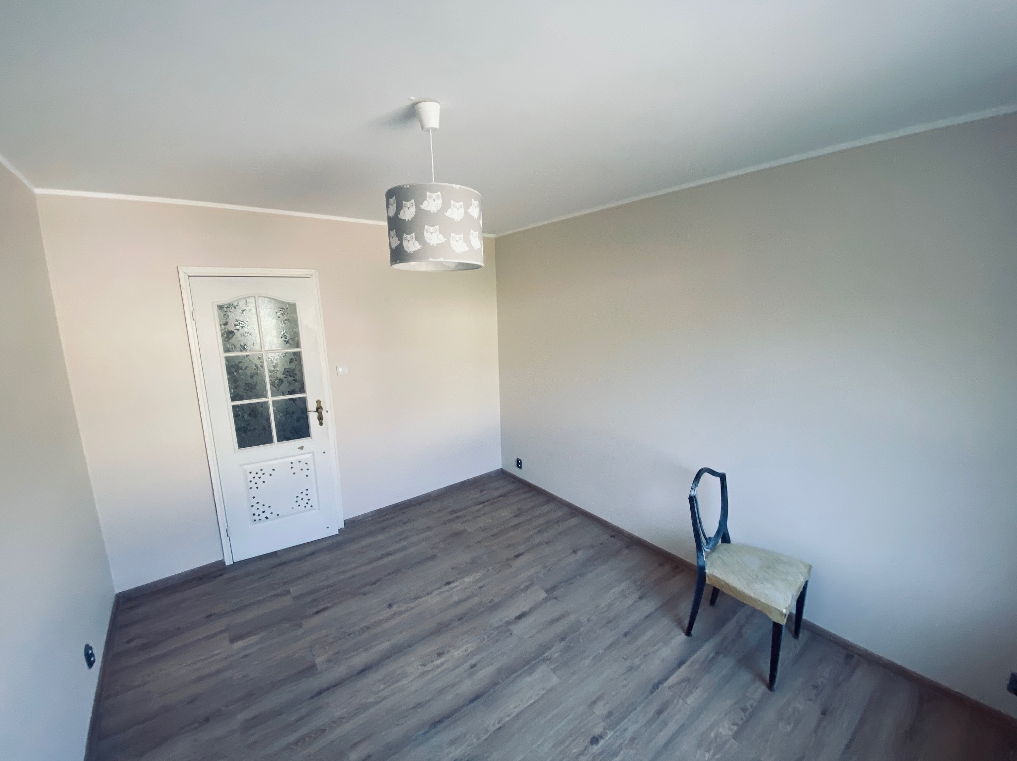 Pomalowane na beżowo wnętrze pokoju z panelami podłogowymi. Widoczny biały sufit, drzwi z szybą i lampa z motywem sów. Stare krzesło przy ścianie.