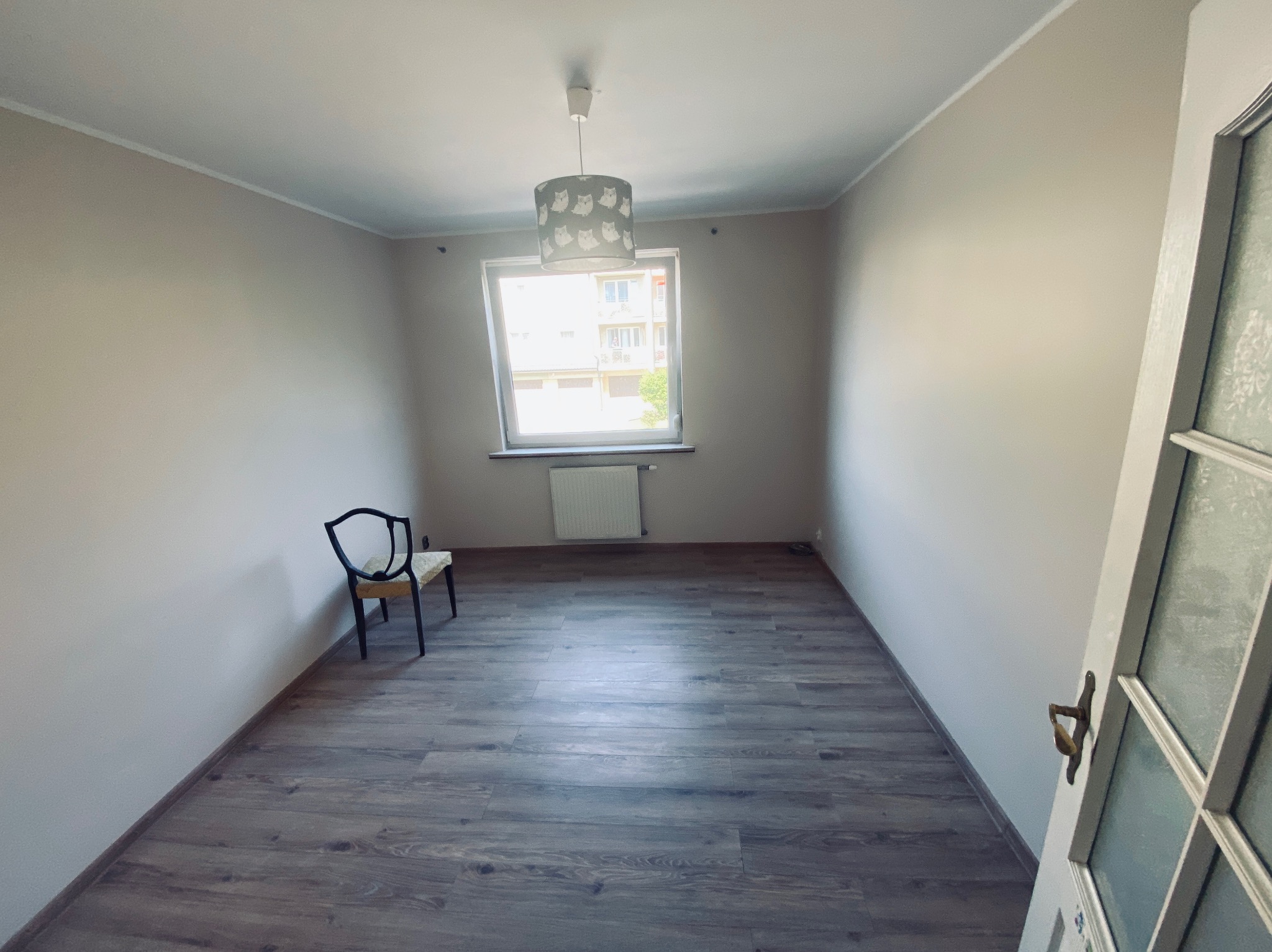 Pusty pokój po remoncie: świeżo ułożone panele podłogowe w odcieniach szarości, pomalowane ściany na jasny beż. W rogu stoi krzesło, a przez okno widać budynek.