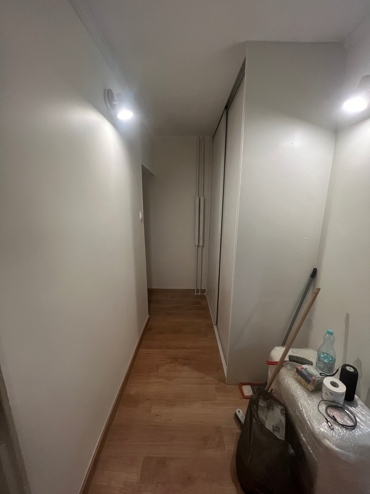 Wnętrze korytarza po remoncie: gładkie, białe ściany z zamontowanym oświetleniem, drewnopodobne panele podłogowe. W tle widoczne drzwi przesuwne szafy.