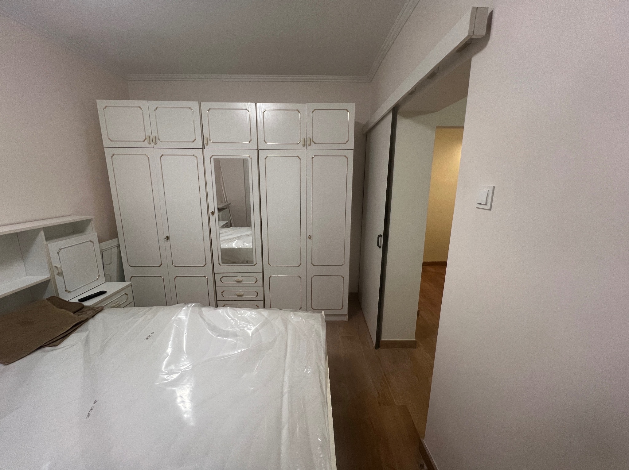 Jasna sypialnia z dużym, białym, ozdobnym zestawem mebli, w tym szafa z lustrem. Podłoga z paneli, ściany w odcieniach beżu. Widoczne drzwi przesuwne.