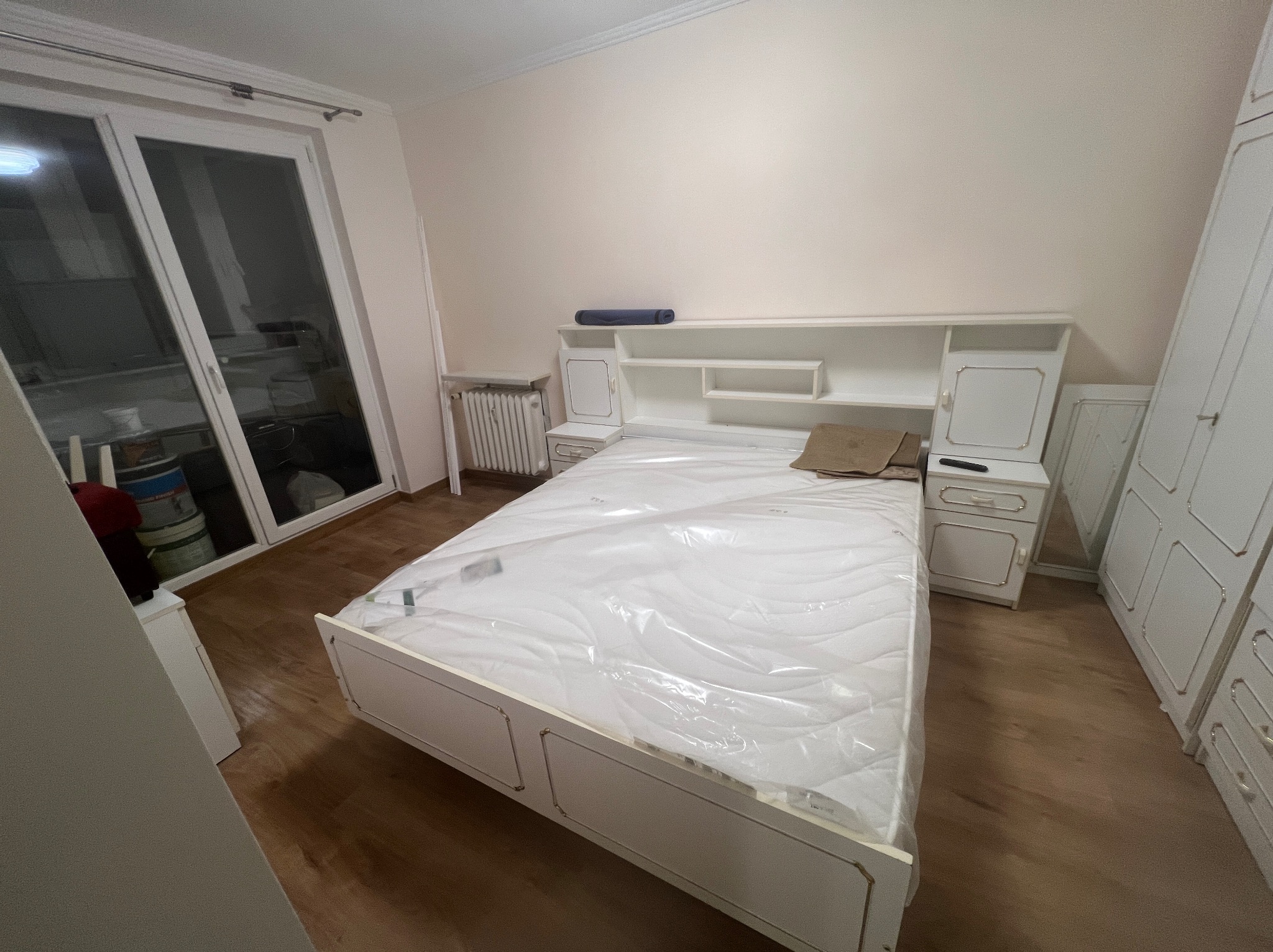 Jasne wnętrze sypialni po wykończeniu: białe meble z dekoracyjnymi detalami, łóżko z materacem w folii, okno balkonowe i parkiet. Styl klasyczny.