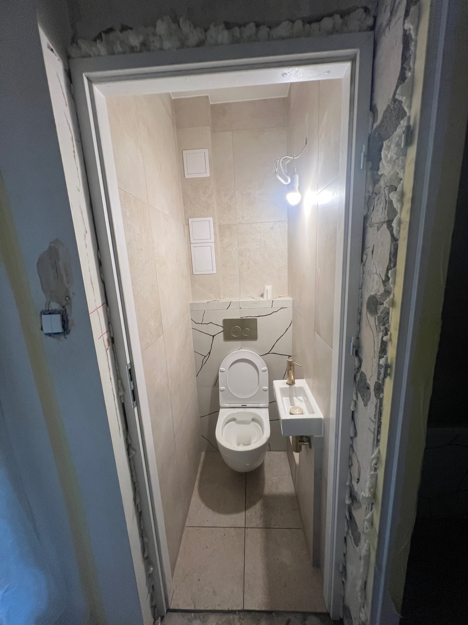 Wykończona łazienka z beżowymi płytkami i białą armaturą, widoczna przez futrynę drzwi w trakcie remontu. Fragment instalacji elektrycznej i surowa ściana w tle.