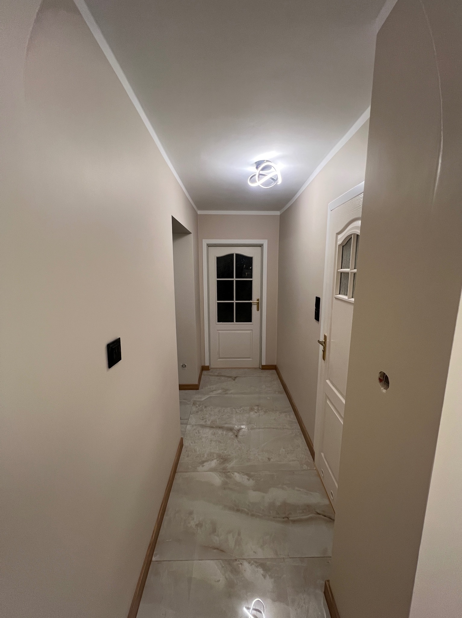 Korytarz po remoncie: beżowe ściany z czarnymi włącznikami, białe drzwi z szybami, marmurowe płytki podłogowe i nowoczesna lampa sufitowa.