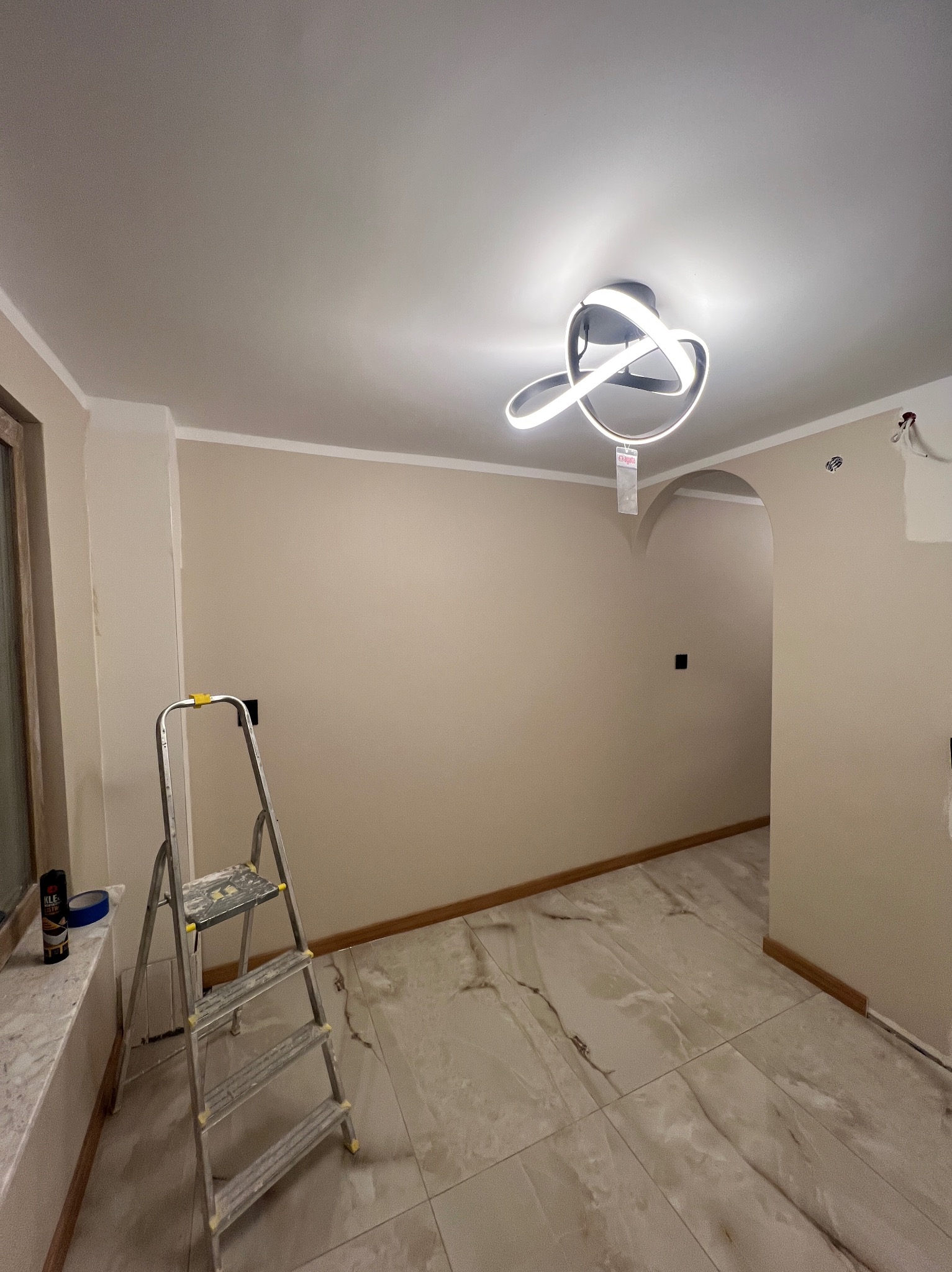 Jasne wnętrze po odświeżeniu: beżowe ściany z listwami, marmurowe płytki, nowoczesna lampa. Aluminiowa drabina malarska przy oknie. Widoczny fragment przejścia.