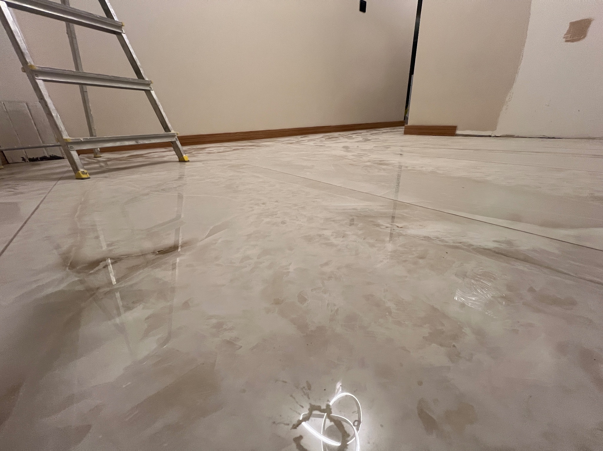 Błyszcząca, beżowa podłoga z płytek imitujących marmur, częściowo zakryta folią ochronną. W tle drabina malarska i ściana z listwą przypodłogową. Widoczne ślady po pracach wykończeniowych.
