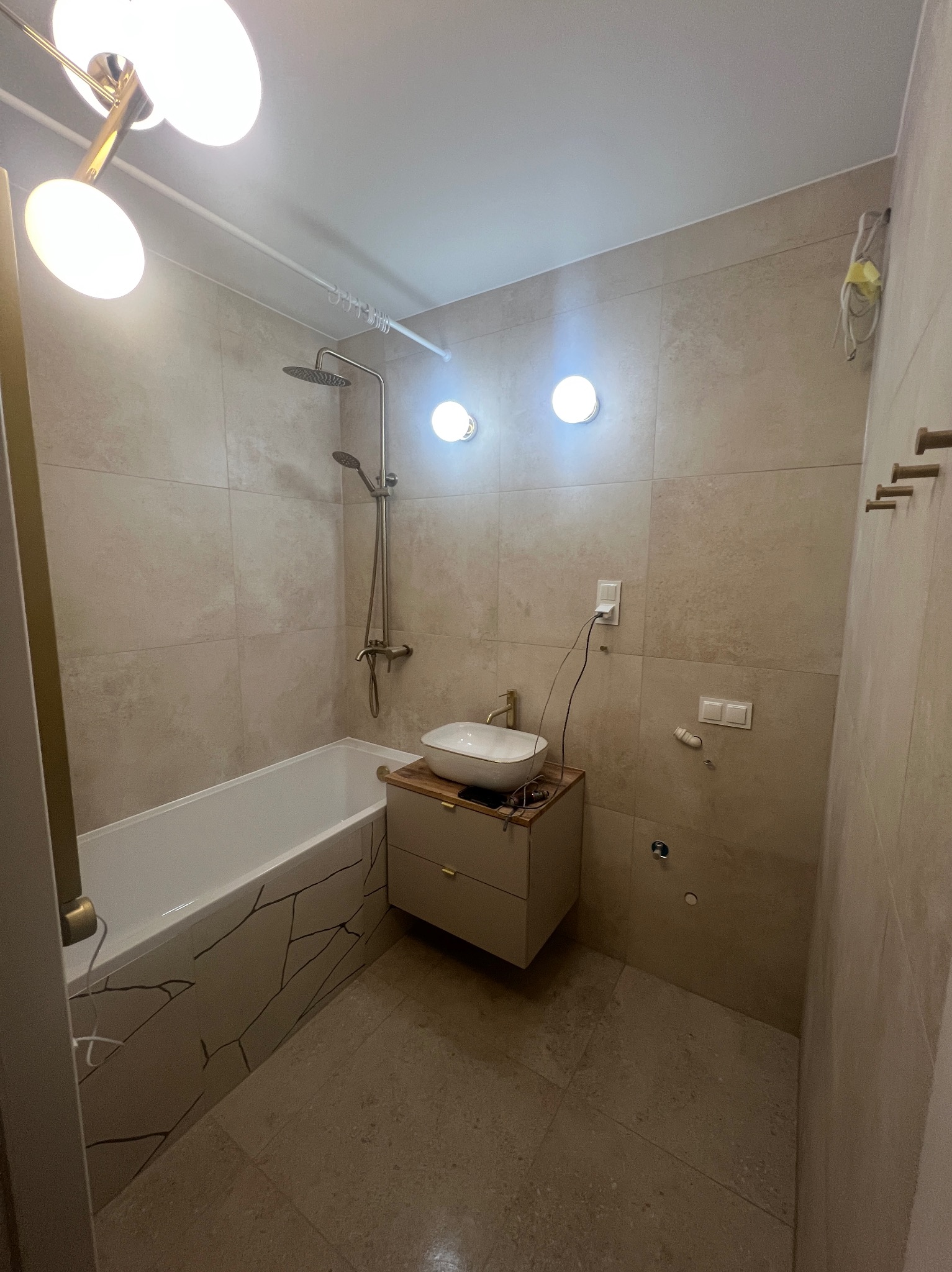 Jasna łazienka z beżowymi płytkami, kabiną prysznicową, umywalką na szafce i wanną z dekoracyjnym wzorem. Widoczne oświetlenie sufitowe i akcesoria łazienkowe.