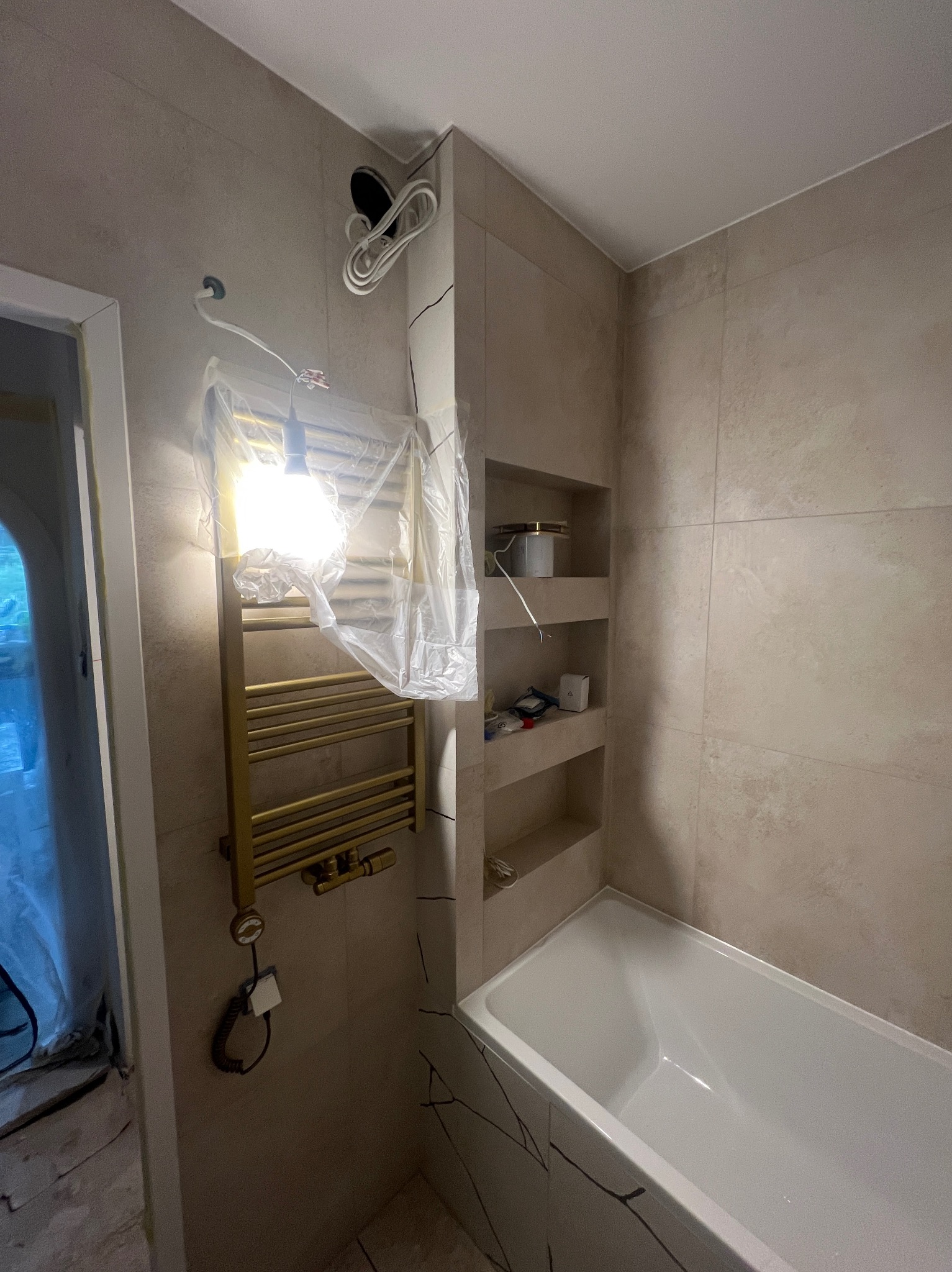 Wykończona łazienka z beżowymi płytkami, wanną i grzejnikiem drabinkowym w trakcie montażu, zabezpieczonym folią. Widoczne półki w ścianie i niedokończone podłogi.
