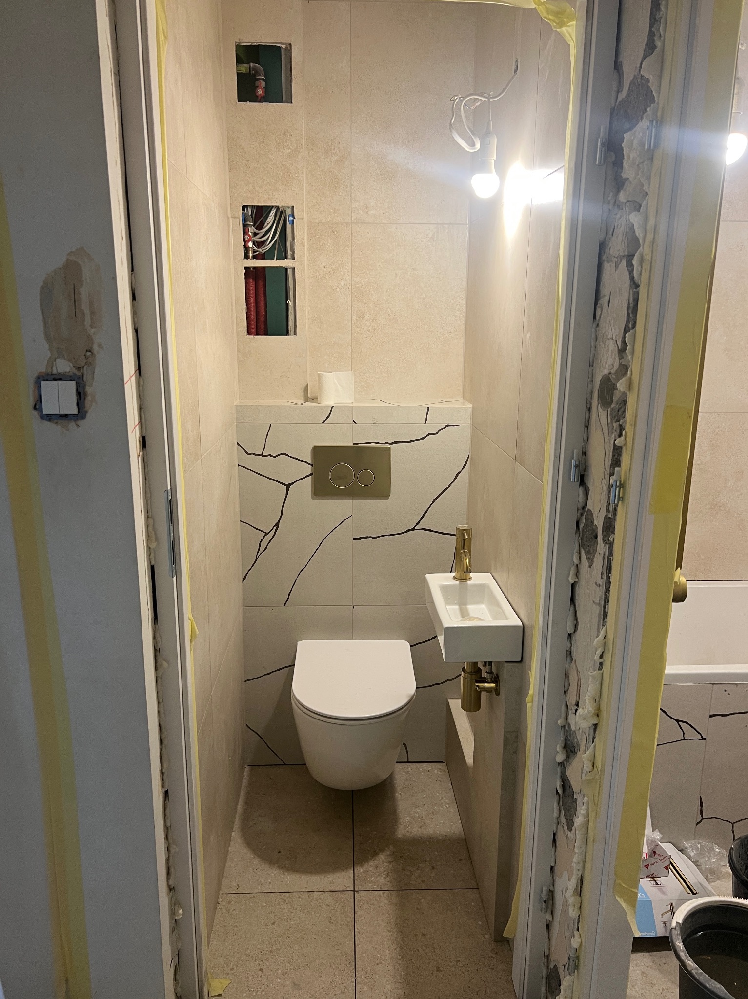 Wykończona łazienka z beżowymi płytkami imitującymi pęknięcia, białą toaletą i umywalką. Widoczne zabezpieczenia taśmą malarską na ościeżnicach.