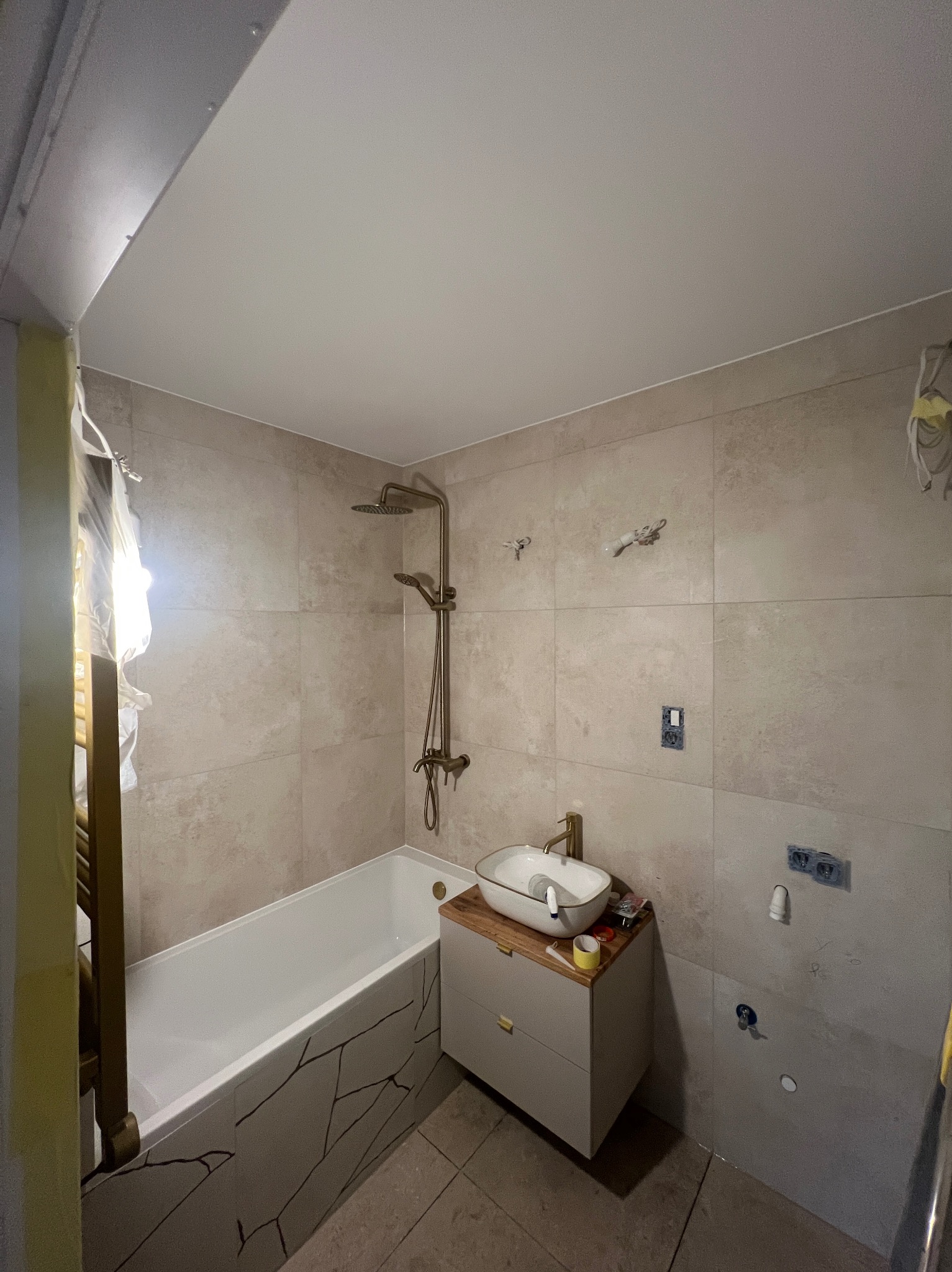 Wykończona łazienka z beżowymi płytkami, białą wanną z dekoracyjnym wzorem i umywalką na szafce. Widoczne elementy instalacji elektrycznej i hydraulicznej.