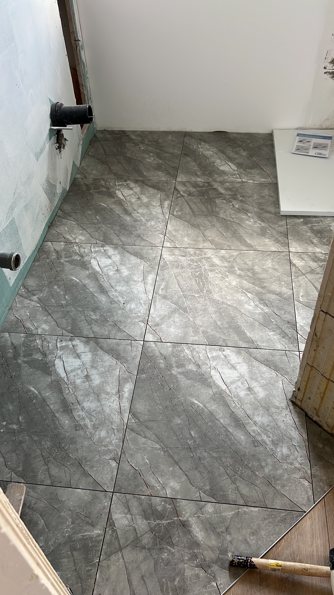 Ułożone szare płytki imitujące marmur w łazience w Szczecinie. Widoczne rury przy ścianie i fragment progu drzwi. Na podłodze leży młotek.