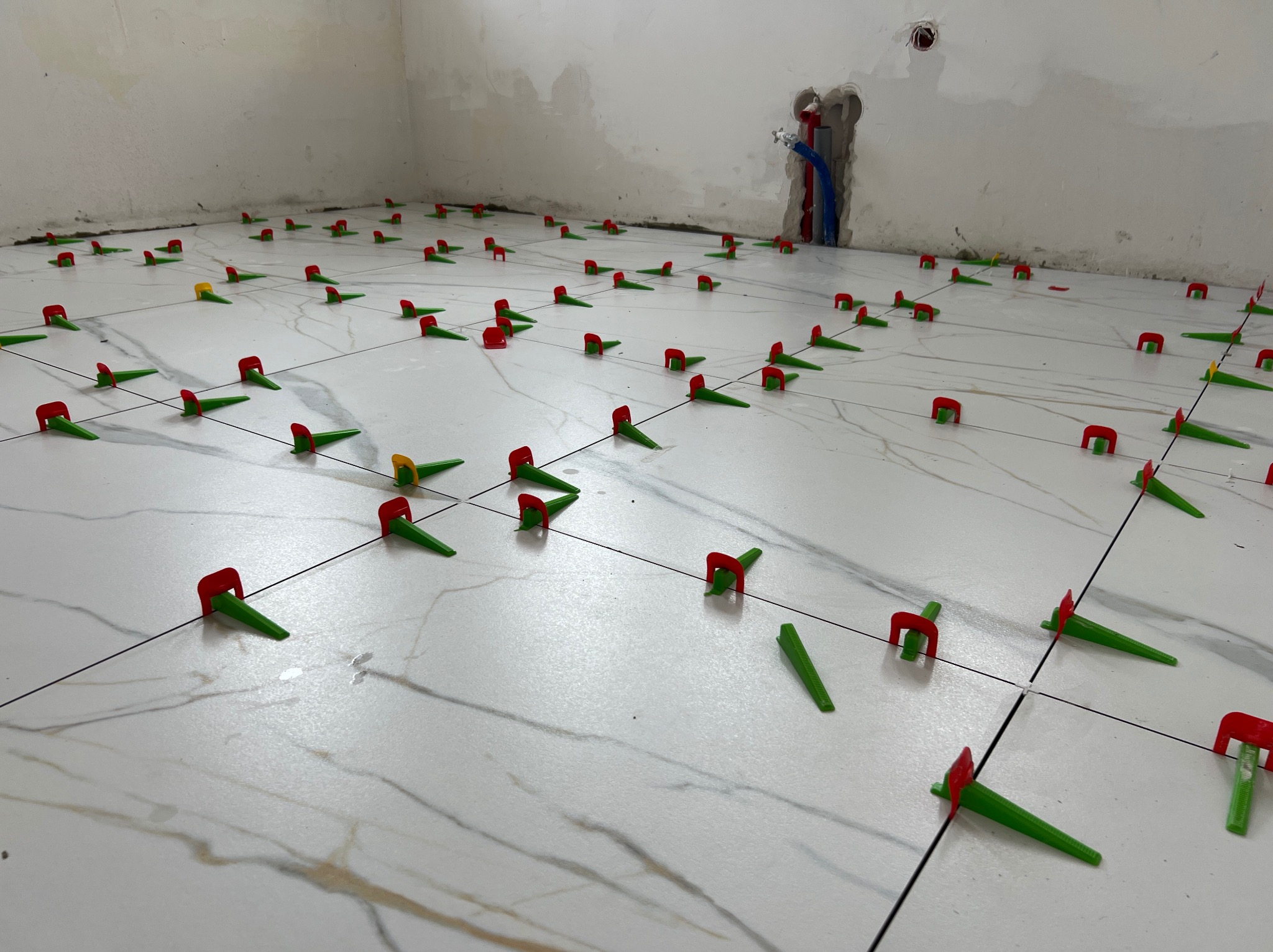 Białe płytki podłogowe z marmurowym wzorem, wyrównane za pomocą klipsów i klinów poziomujących. W tle ściana z widocznymi śladami po pracach instalacyjnych.