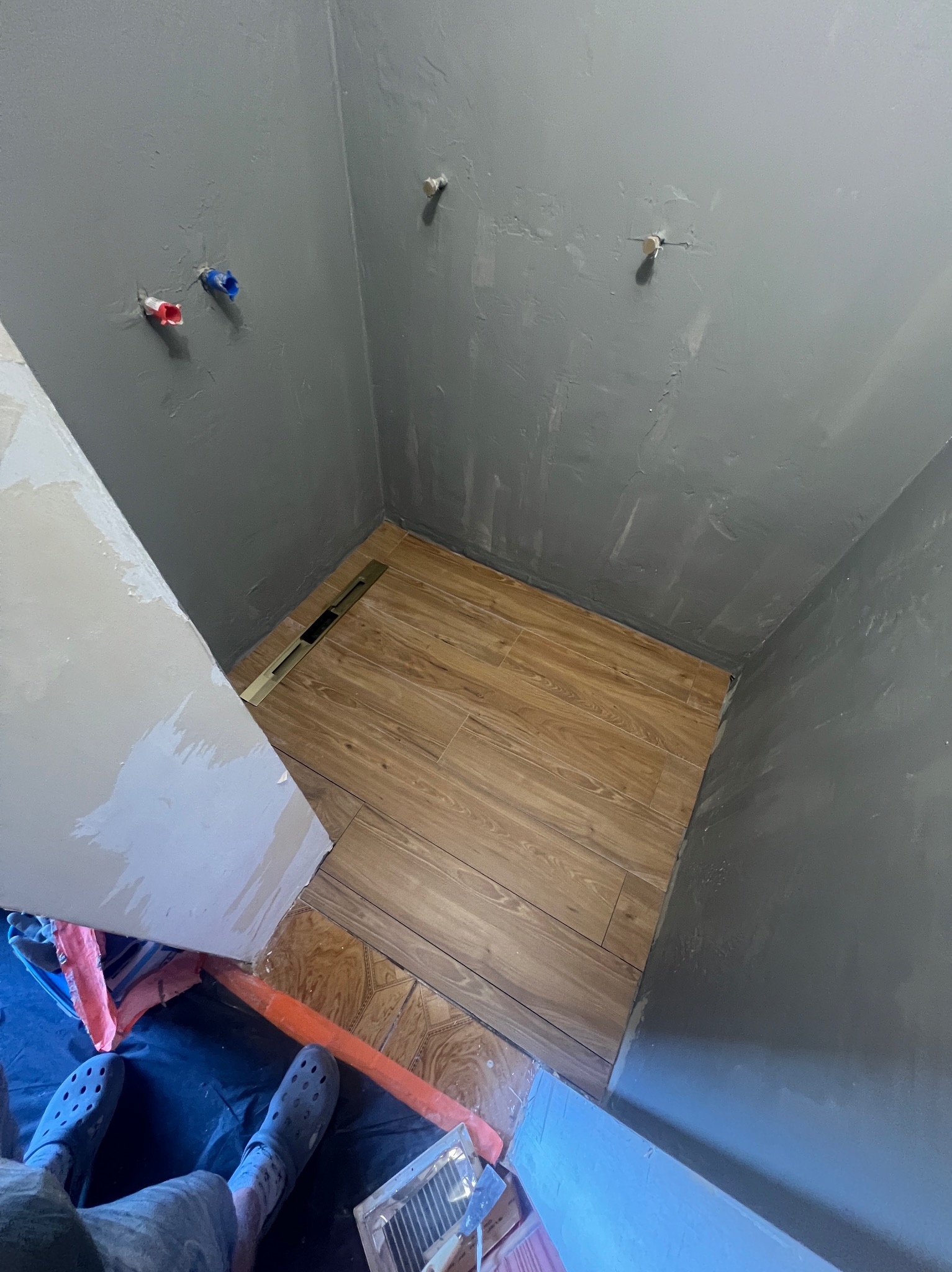 Szare ściany łazienki z instalacją wodną, drewnopodobne płytki podłogowe z odpływem liniowym. Widoczne narzędzia i fragment osoby w obuwiu roboczym.