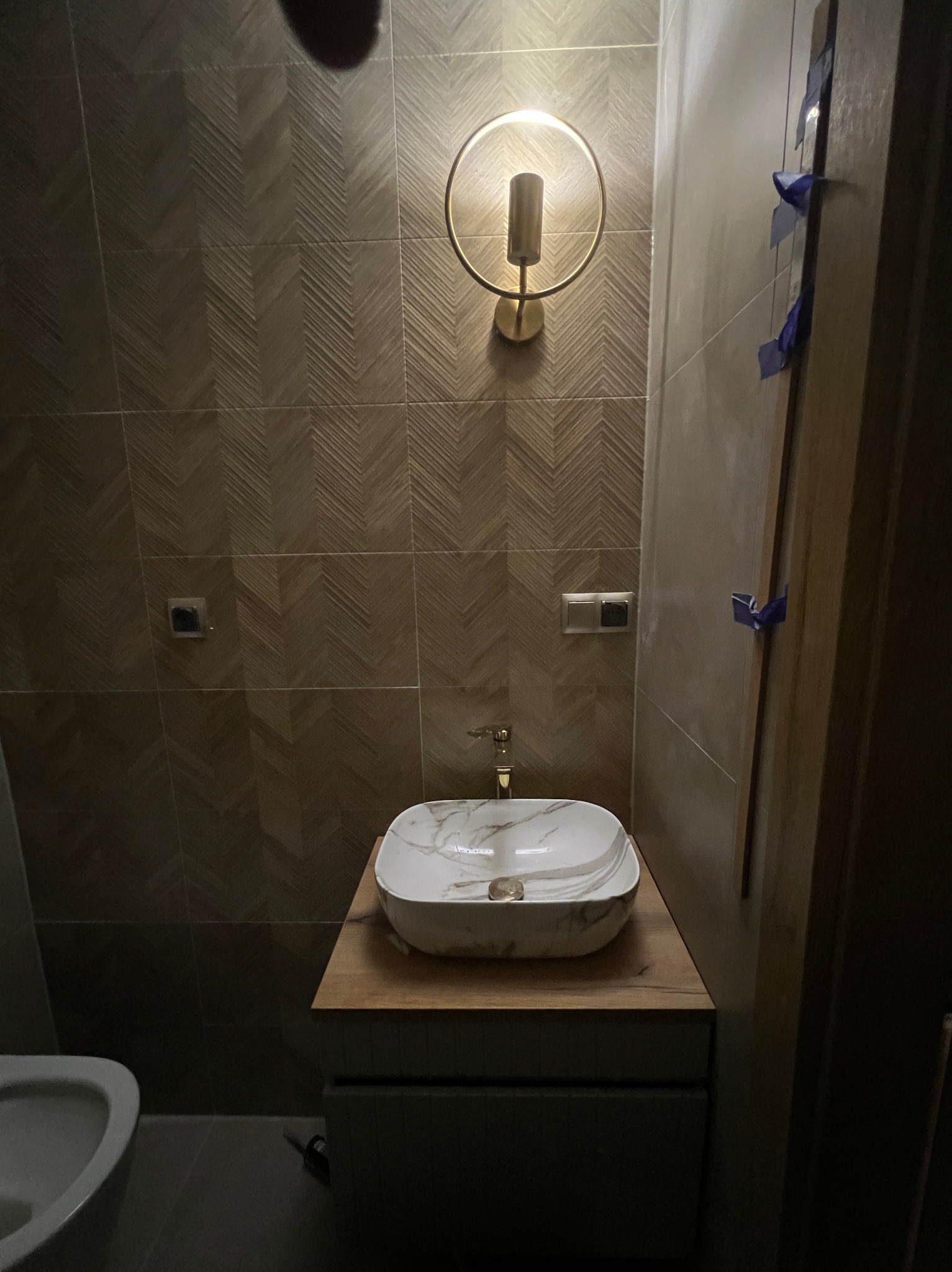 Wykończona łazienka z umywalką w stylu marmuru, drewnianym blatem i złotą lampą na ścianie z płytek w jodełkę. Widoczne detale wykończeniowe.