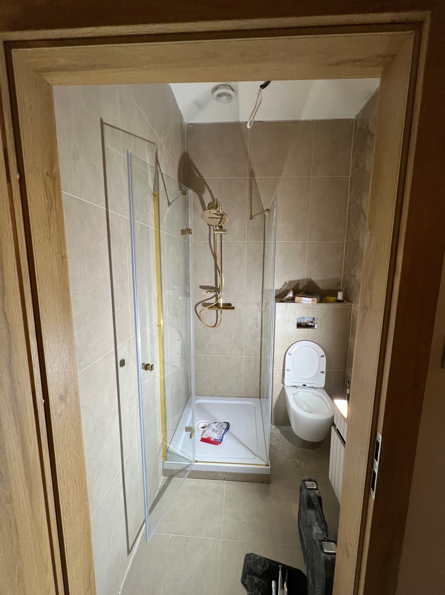 Wykończona łazienka z kabiną prysznicową ze złotymi elementami, toaletą i beżowymi płytkami. Widok zza drewnianej futryny drzwi.