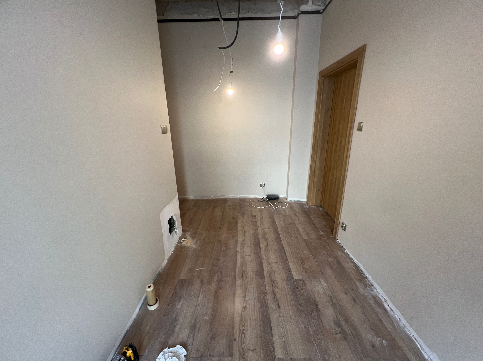Wnętrze po remoncie: świeżo ułożone panele podłogowe w odcieniach brązu, pomalowane na beżowo ściany, widoczne kable i żarówki, drewniane drzwi.