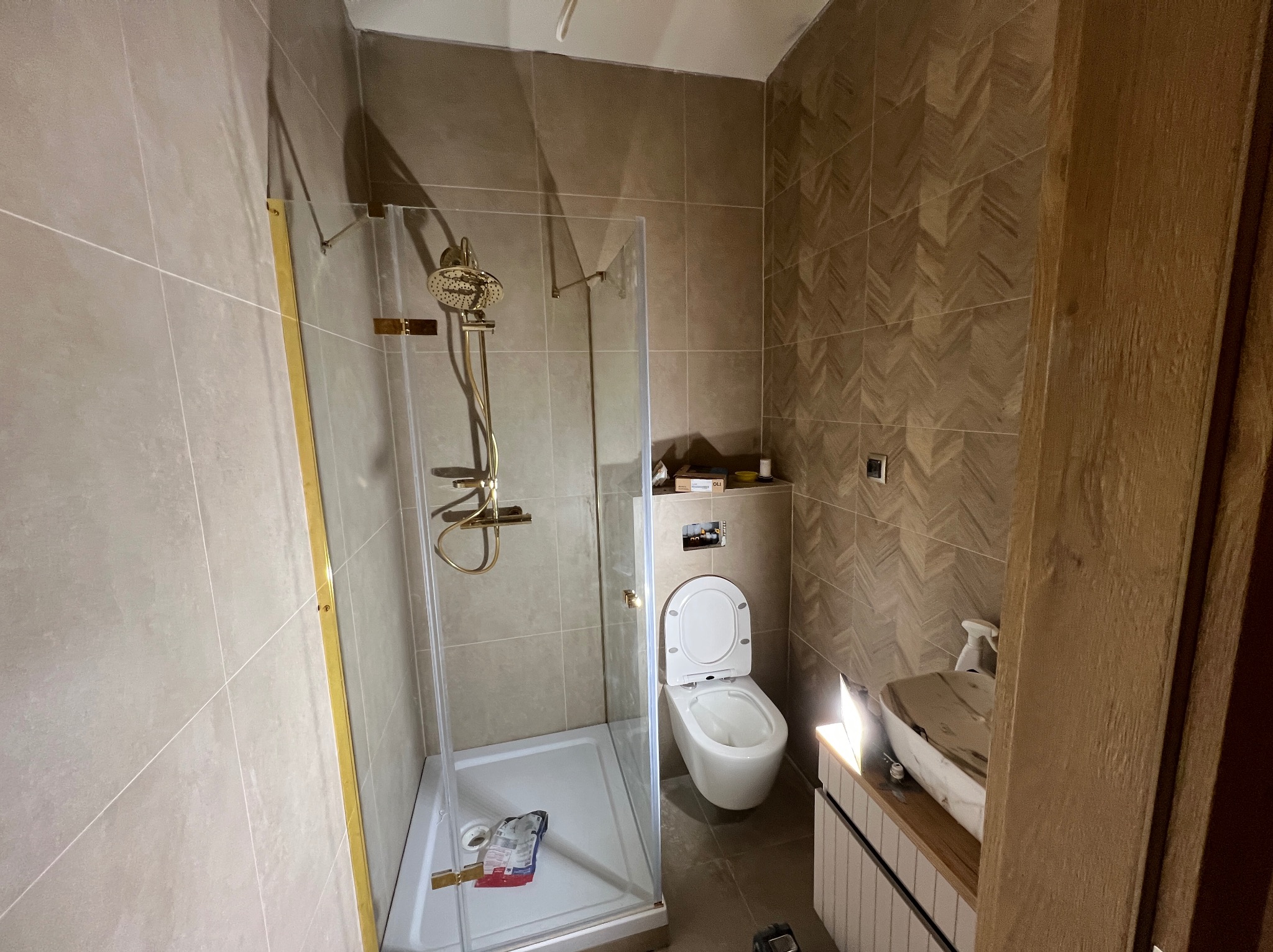 Wykończona łazienka z kabiną prysznicową ze złotymi elementami, toaletą podwieszaną i umywalką nablatową. Na ścianach płytki w odcieniach beżu i brązu.