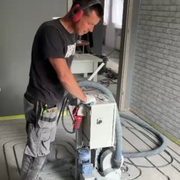 Feniks Damian Piec - Pracownik w słuchawkach szlifuje posadzkę z ogrzewaniem podłogowym za pomocą specjalistycznej maszyny. Widoczne rury ogrzewania w podłodze i ściana z cegły.