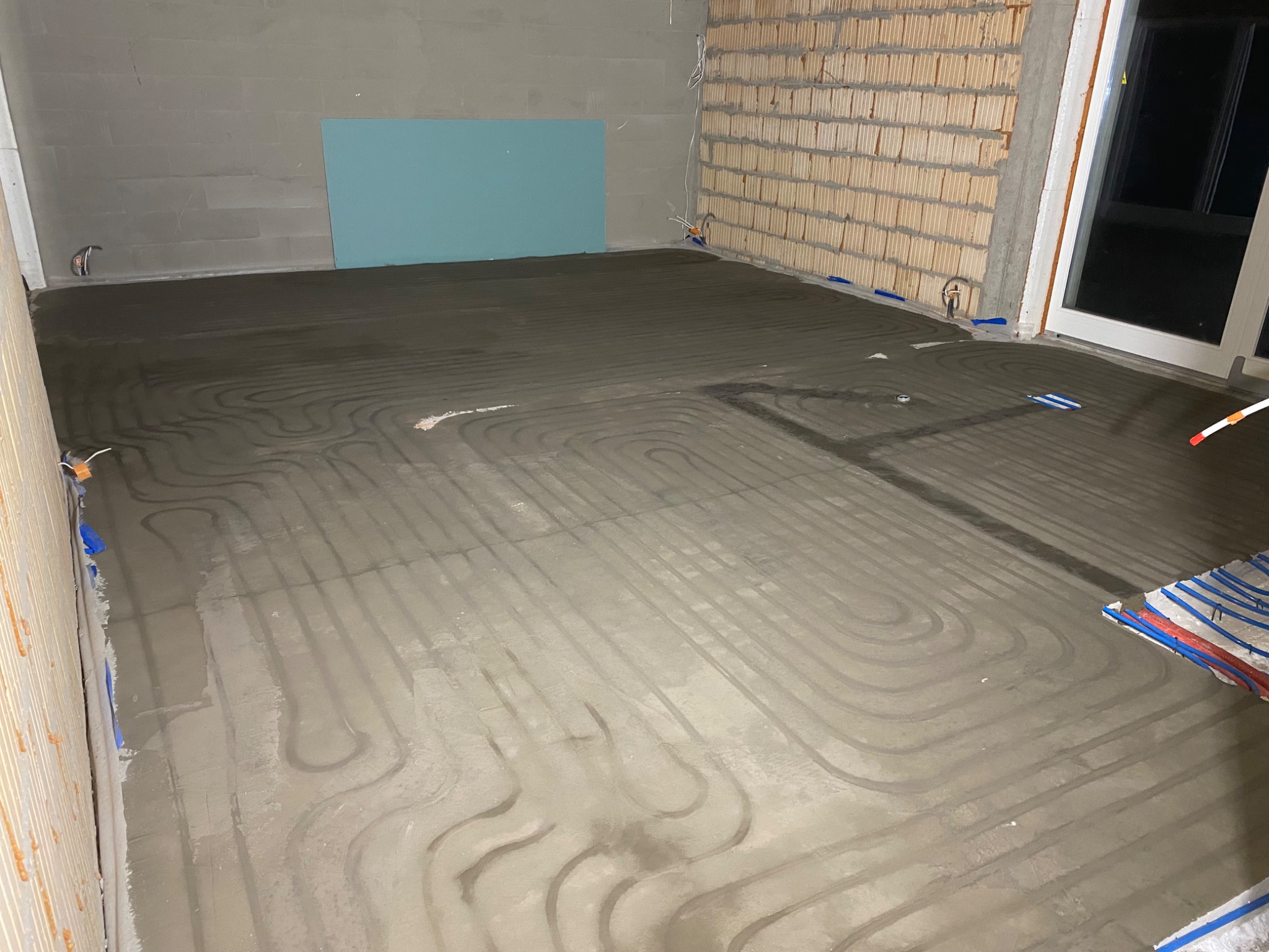 Szare rury ogrzewania podłogowego zalane wylewką, widoczne fragmenty ścian z cegły i bloczków, niebieska płyta gipsowa. Instalacja w trakcie realizacji.