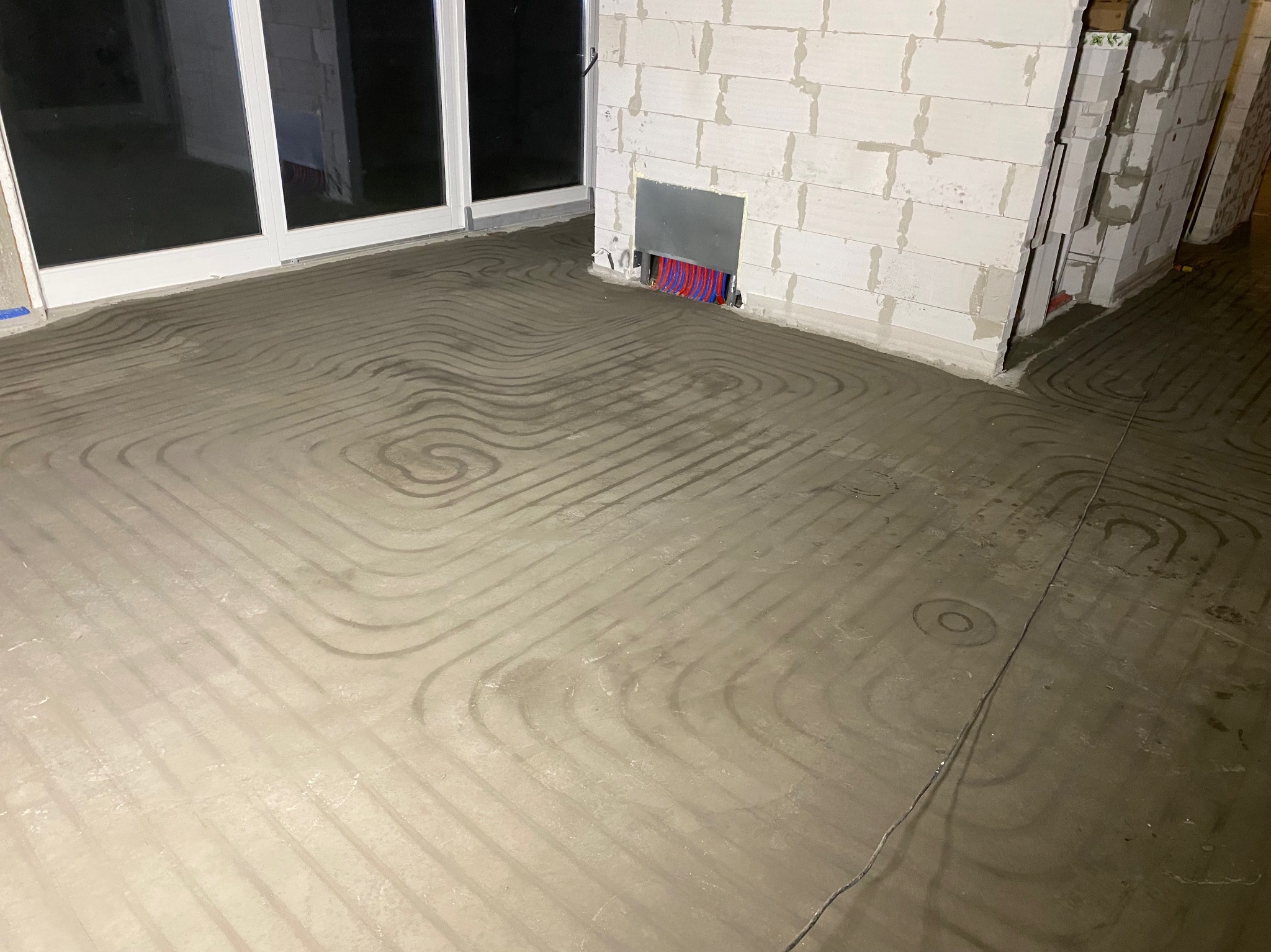 Zalana posadzka z widocznym układem rur ogrzewania podłogowego w surowym wnętrzu. Fragment ściany z białych bloczków i okno balkonowe w tle.