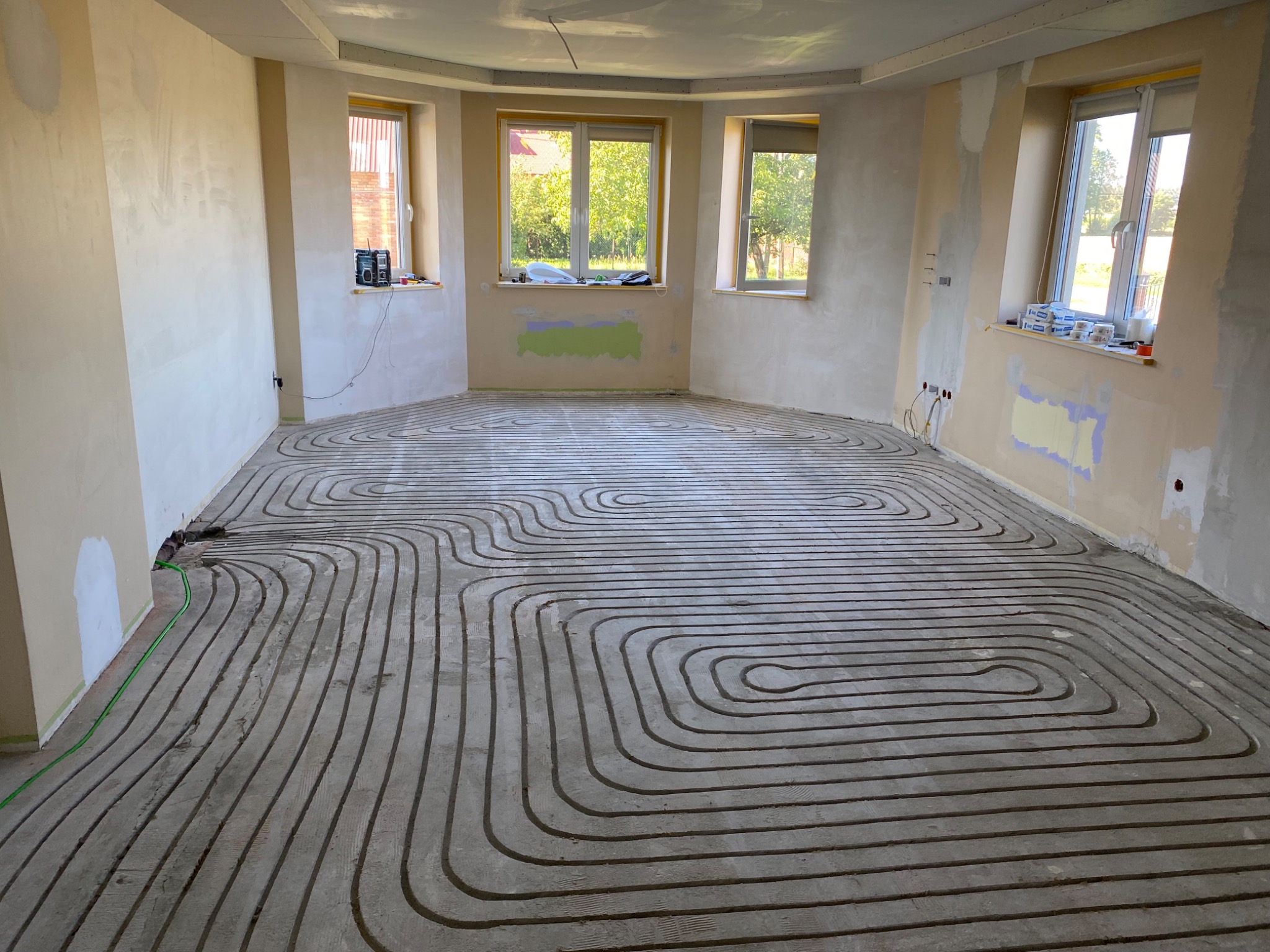 Szare rurki ogrzewania podłogowego ułożone w zygzak na podłodze w surowym pomieszczeniu z trzema oknami. Widoczne ślady po łatach na ścianach.