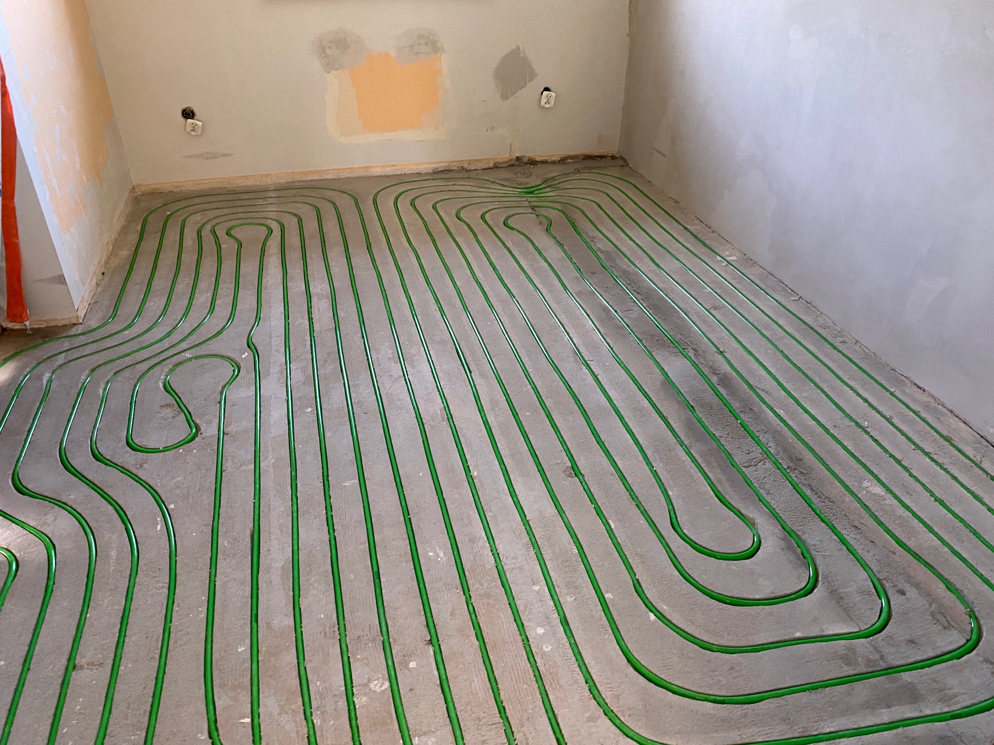 Zielone rury ogrzewania podłogowego ułożone w pętle na szarej, betonowej posadzce w surowym wnętrzu. Widoczne ślady po łatach na ścianie.